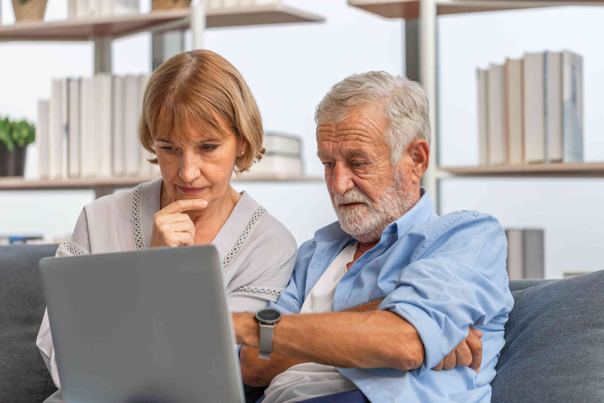 An elderly couple sits on a couch, looking at their laptop screen as if searching for something.